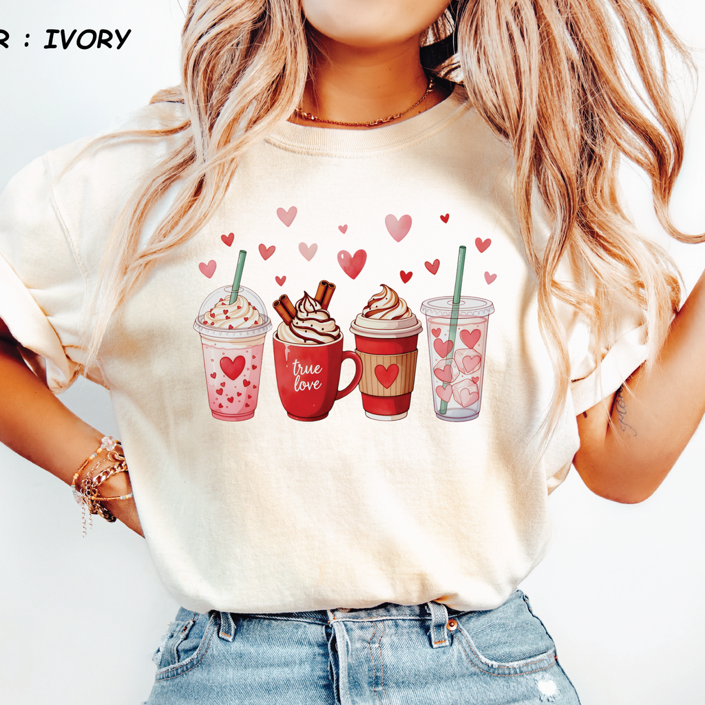 Ivory Comfort Colors t-shirt featuring a watercolor illustration of four Valentine's Day coffee drinks including a pink frappe and iced coffee.