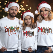 Family wearing matching Christmas sweatshirts reading “Daddy Claus”, “Mama Claus”, “Mini Claus”, “Granny Claus” with festive plaid and candy-cane letters